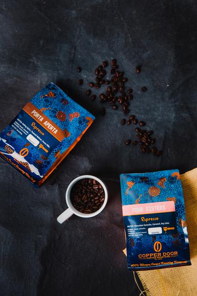 Two bags of Copper Door Coffee Roasters coffee beans with a mug and beans around them
